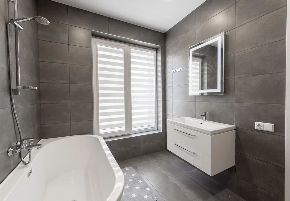 Intérieur d'une salle de bain en rénovation montrant deux artisans en action : un électricien en tenue professionnelle travaillant sur des installations électriques (tableautin, câblage) en hauteur ou sur un mur, et un plombier en tenue professionnelle installant ou réparant des éléments sanitaires (lavabo, toilettes) simultanément dans le même espace. Les deux professionnels sont visibles et impliqués dans leurs tâches respectives, illustrant clairement la collaboration et la double compétence sur un même chantier de rénovation intérieure.