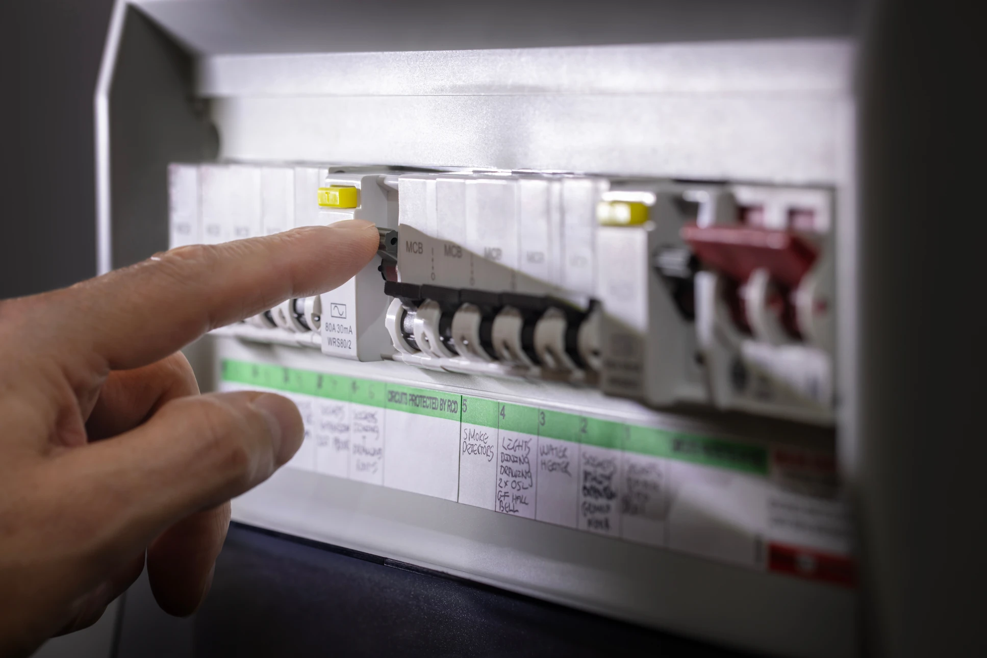 Photographie réelle en couleur montrant un électricien professionnel effectuant une mise aux normes électrique dans un intérieur résidentiel. L'électricien, vêtu de façon professionnelle, manipule des câbles électriques ou travaille sur un tableau électrique normalisé, dans un contexte d'habitation (murs, plinthe, luminaire visible). L'image doit transmettre la professionnalité, la rigueur technique et l'assurance de la conformité aux normes de sécurité électrique.